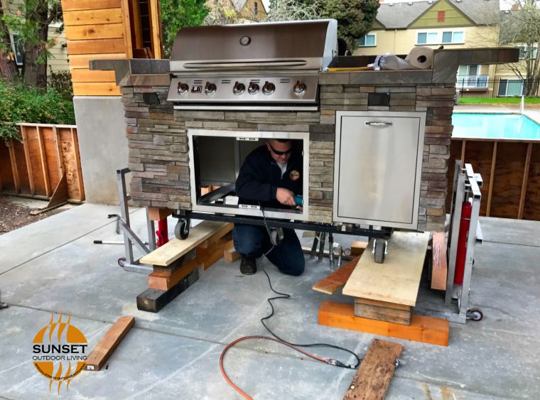 outdoor kitchen Install by sunset outdoor living stainless steel appliances with earth tone small stone brick up on blocks while installer is inside